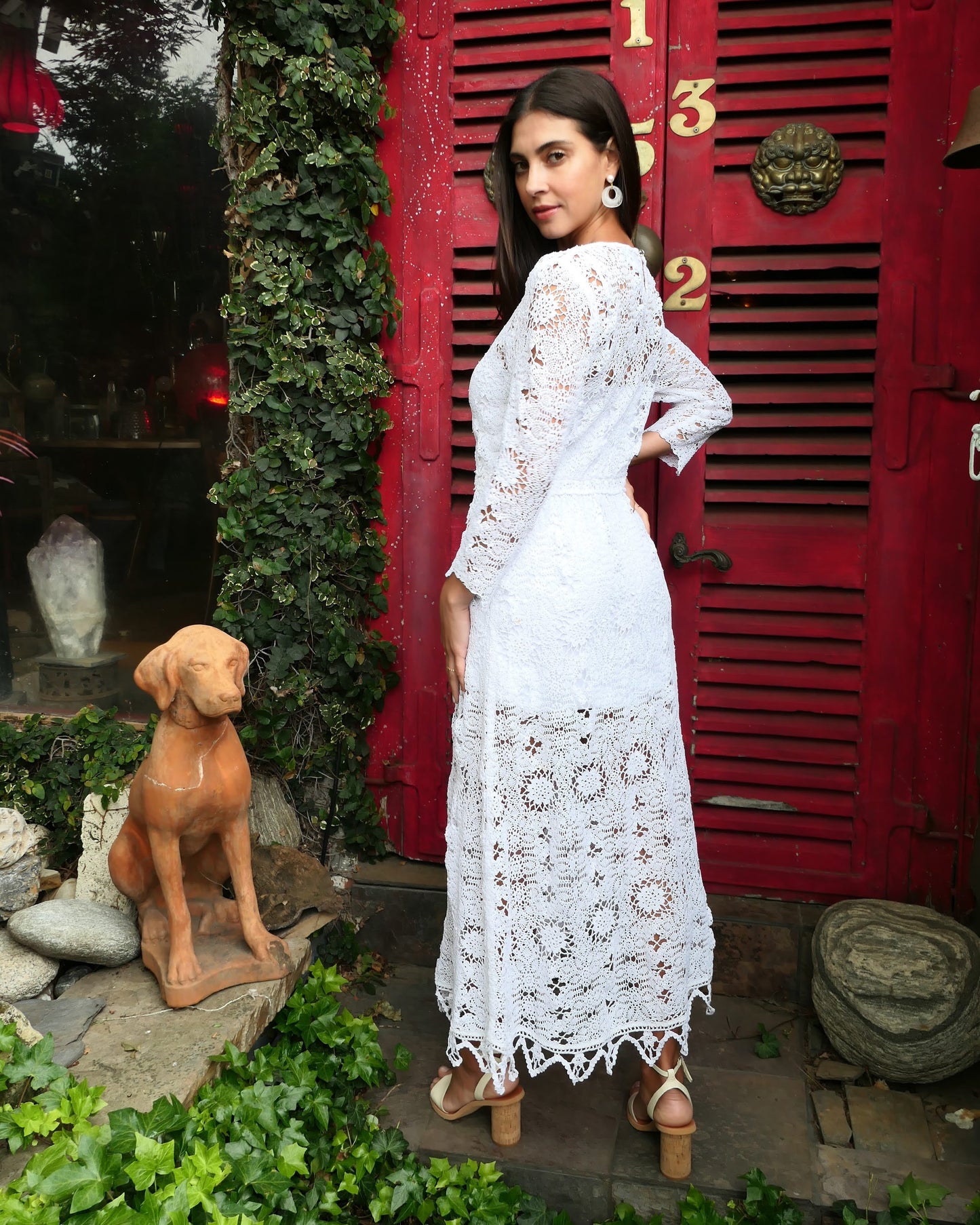 Back view of model wearing dress. A maxi dress with a classic silhouette, long sleeves, and a comfortable elastic waistband. Stitched together by hand using individual vintage doily lace pieces. Throw it over a white slip and a pair of open-toe heels for a stroll out on the town at your favorite beach destination. Color: White