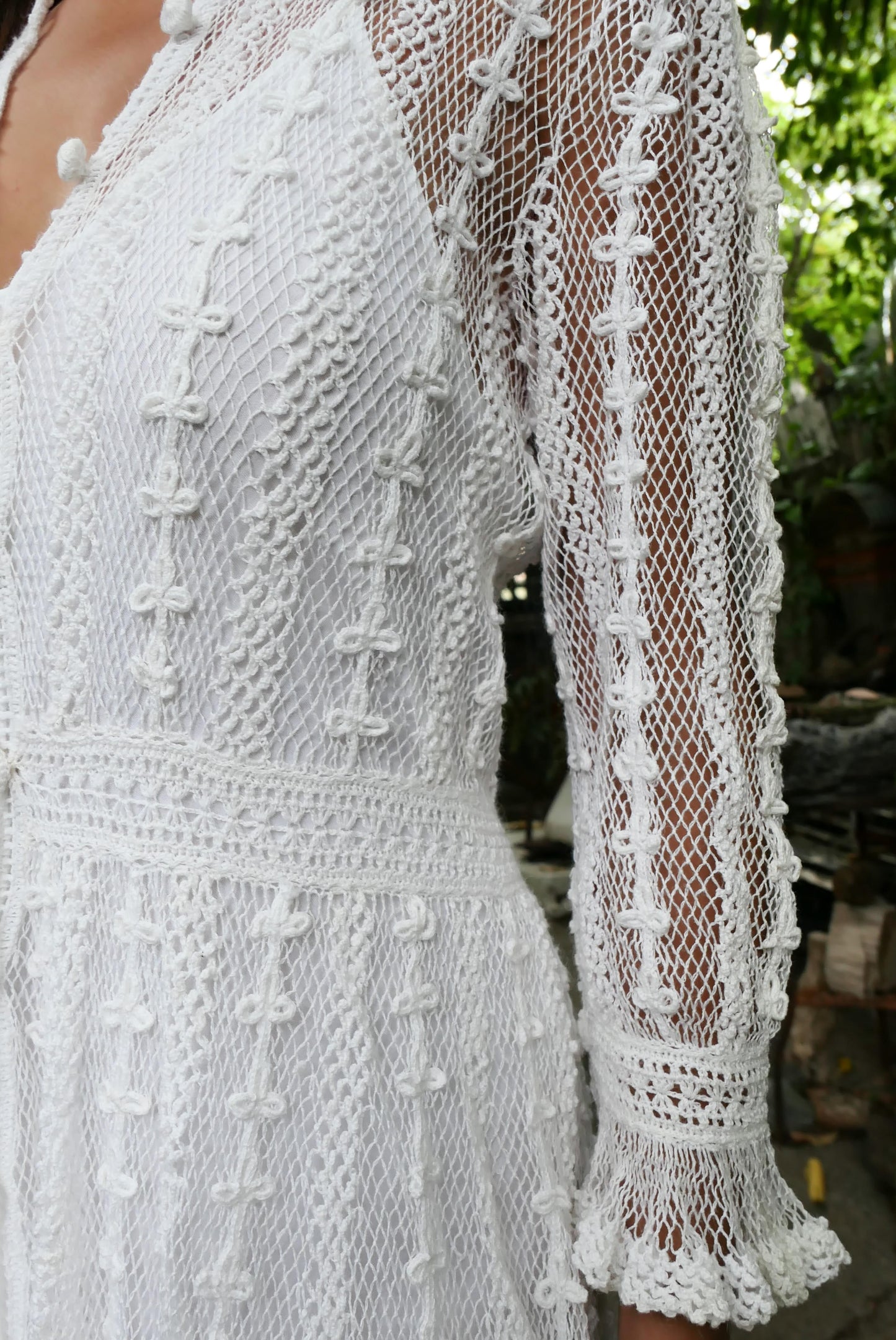 Closeup of sleeve. Ruffles out from the wrist. Lim's Vintage original crochet cottage core style dress. This dreamy midi length dress can be worn buttoned up the front or back and is made with very fine threads, making the dress refined and sheer. Detailed crochet trim is used for the waist, hemline, and 3/4 length ruffled sleeves. Wear it with a natural or pink colored slip or bandeau bra and bikini shorts and a pair of ankle or knee-high boots to complete the look.