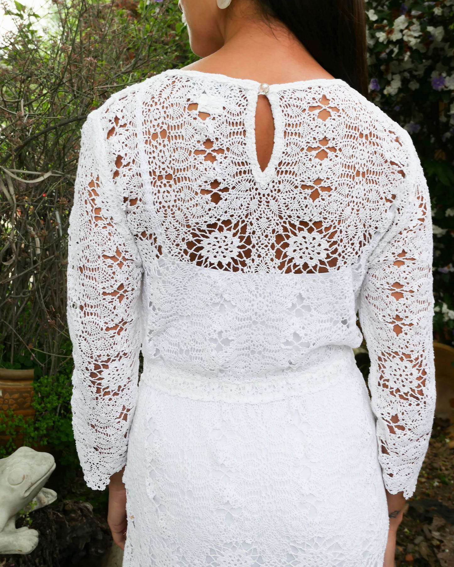 Closeup of back of dress. A maxi dress with a classic silhouette, long sleeves, and a comfortable elastic waistband. Stitched together by hand using individual vintage doily lace pieces. Throw it over a white slip and a pair of open-toe heels for a stroll out on the town at your favorite beach destination. Color: White