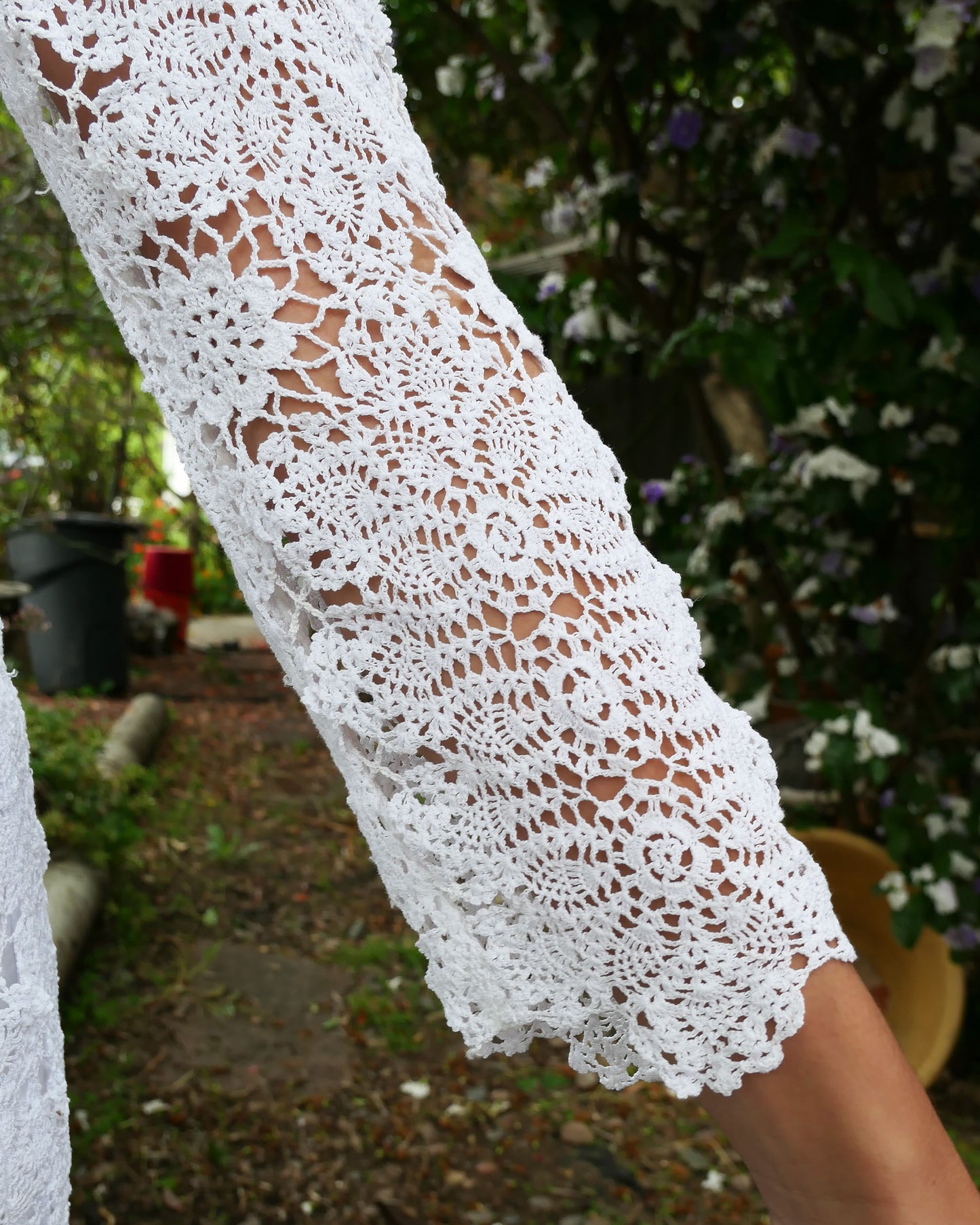 Closeup of sleeve and doily detail. A maxi dress with a classic silhouette, long sleeves, and a comfortable elastic waistband. Stitched together by hand using individual vintage doily lace pieces. Throw it over a white slip and a pair of open-toe heels for a stroll out on the town at your favorite beach destination. Color: White