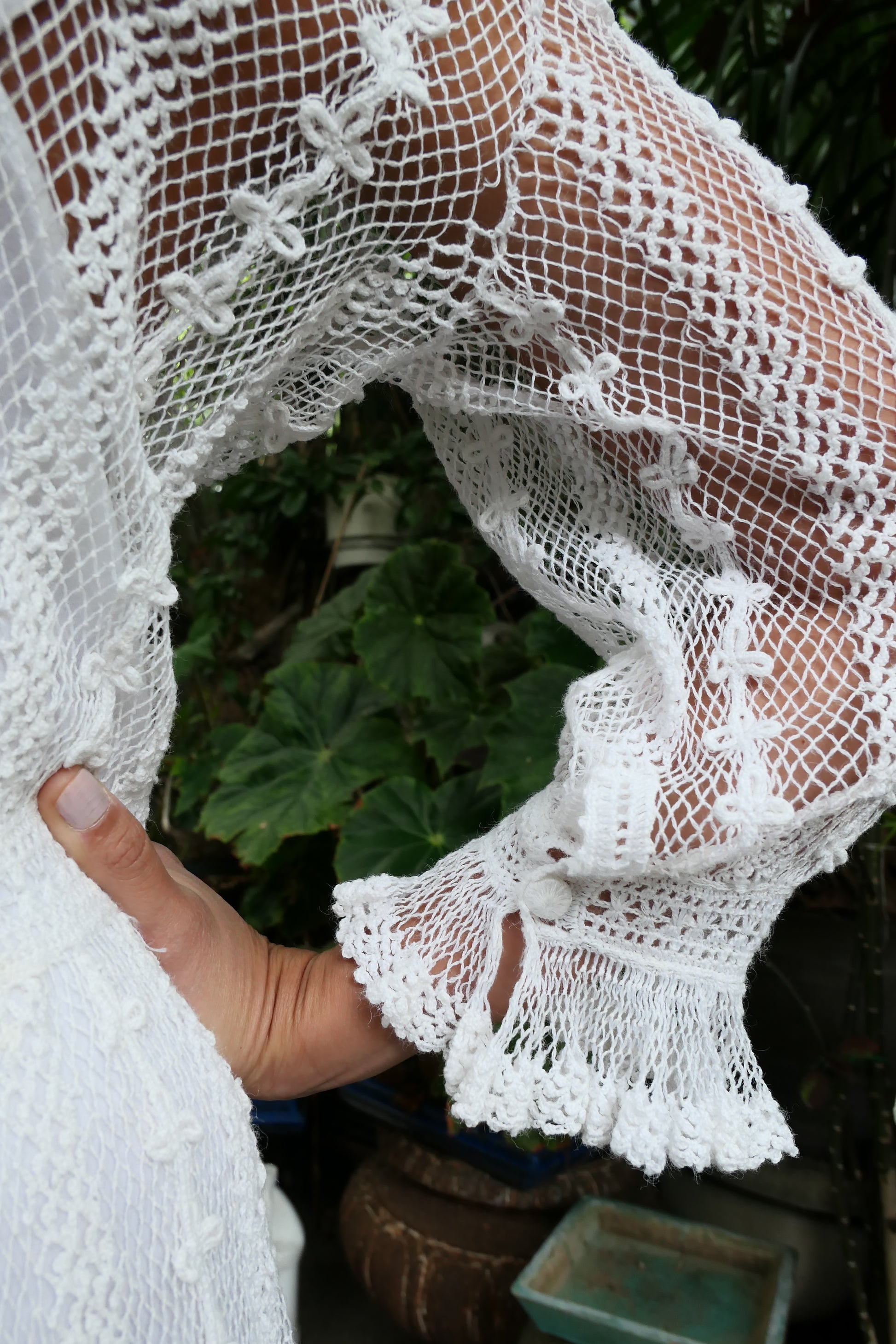 Closeup of sleeve and button-up cuff at the wrist.  Lim's Vintage original crochet cottage core style dress. This dreamy midi length dress can be worn buttoned up the front or back and is made with very fine threads, making the dress refined and sheer. Detailed crochet trim is used for the waist, hemline, and 3/4 length ruffled sleeves. Wear it with a natural or pink colored slip or bandeau bra and bikini shorts and a pair of ankle or knee-high boots to complete the look.