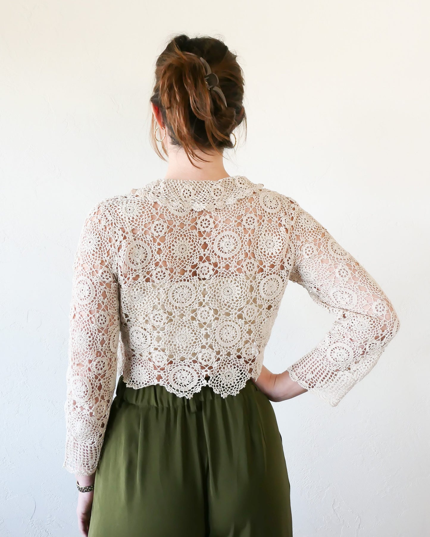 Back view of natural colored crochet cropped cardigan. A versatile and boho chic hand crocheted cropped cardigan sweater with a unique, repeating kaleidoscope-like floral pattern throughout. Dress it down with a pair of jeans and sandals, or dress it up with neutral colored wide leg pants, heels, and a pair of vintage dangly earrings.