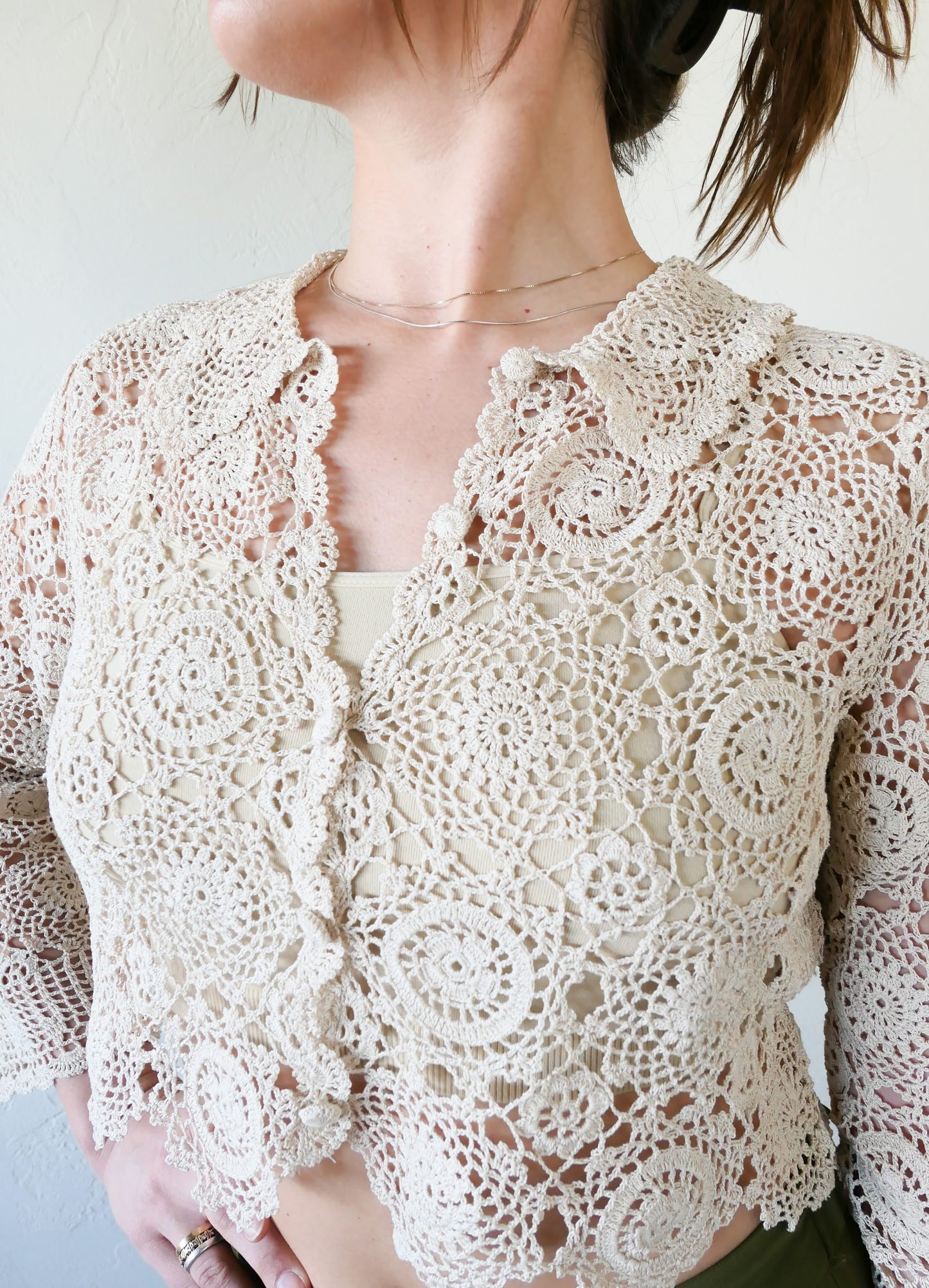 Closeup front view of natural colored crochet crop cardigan. A versatile and boho chic hand crocheted cropped cardigan sweater with a unique, repeating kaleidoscope-like floral pattern throughout. Dress it down with a pair of jeans and sandals, or dress it up with neutral colored wide leg pants, heels, and a pair of vintage dangly earrings.