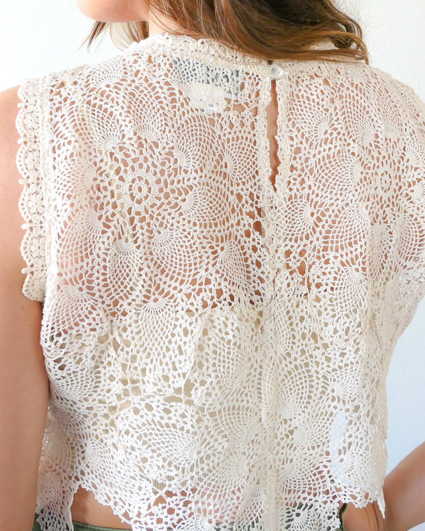 Closeup of back view of top. An ultra casual, hip, and boho-chic crop tank top made with interconnected flower doilies reminiscent of the white lotus, which symbolizes peace, tranquility, and calmness.  Lace trim at the collar and sleeve.  Model is wearing a natural colored crochet top. 