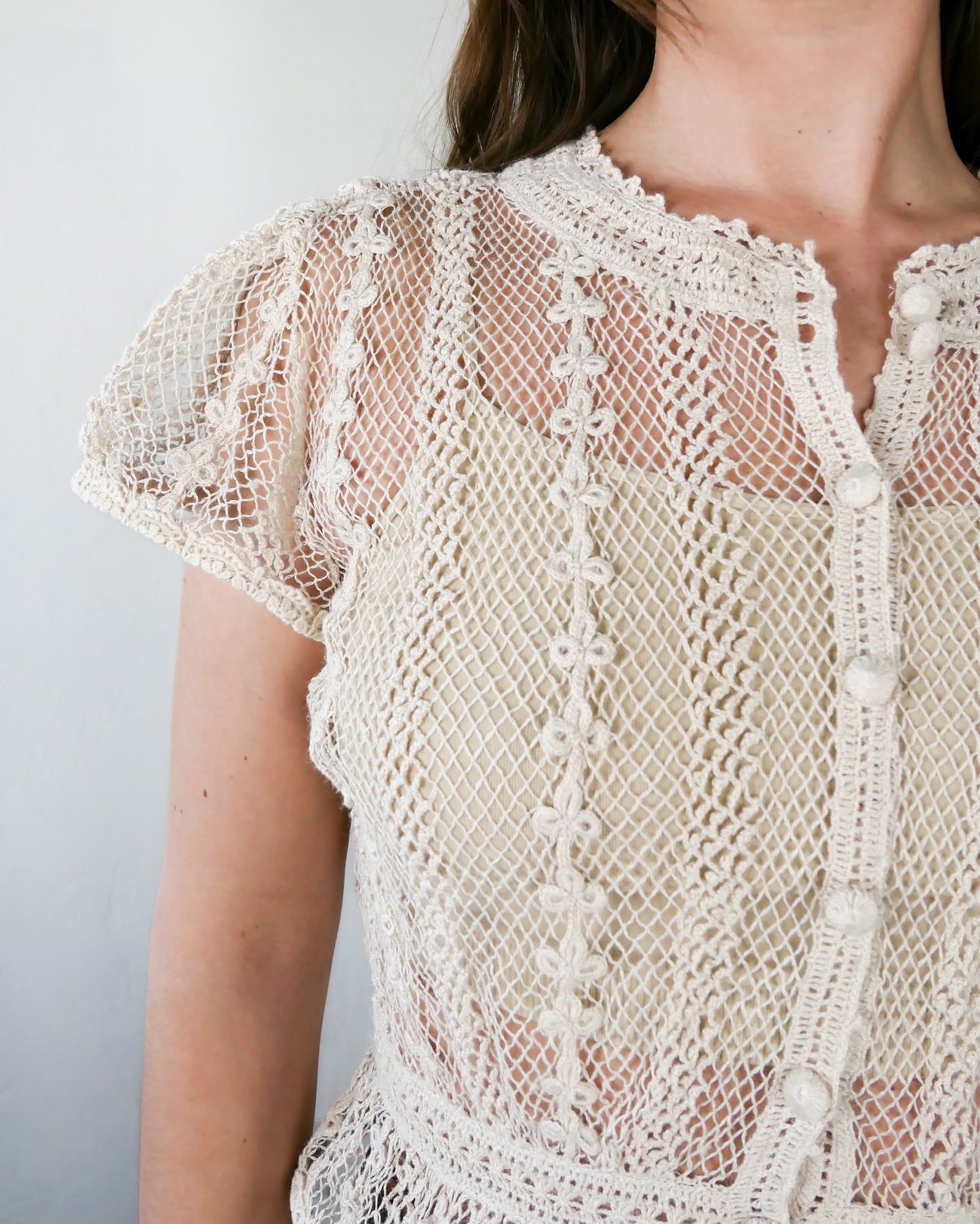 Closeup of front of crochet top in natural color. A Lim's original vintage crochet piece from the 1980s, this top was hand crocheted with very fine threads to create a delicate, lightweight, and transparent feel. Puffed sleeves, a high waist, and flared hem lend the top a whimsical yet tailored look. The top is reversible and can be buttoned up the front or back. Natural color.