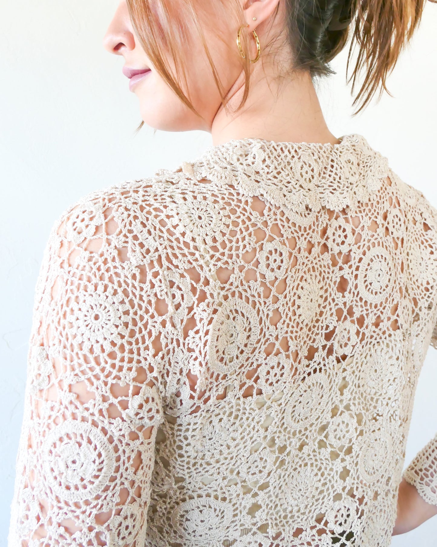Closeup of back side view of collar and upper back. A versatile and boho chic hand crocheted cropped cardigan sweater with a unique, repeating kaleidoscope-like floral pattern throughout. Dress it down with a pair of jeans and sandals, or dress it up with neutral colored wide leg pants, heels, and a pair of vintage dangly earrings.