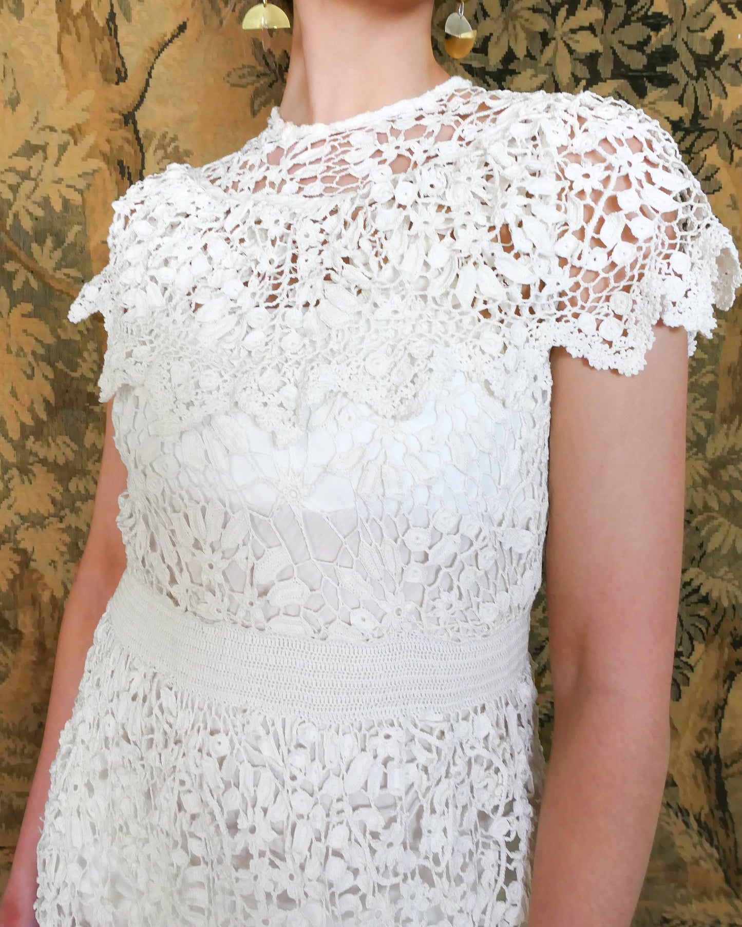 A romantic white crochet midi dress with a floral and leaf pattern, comfortable elastic waistband, and a flattering neck and shoulder design. Perfect as a vintage wedding dress or a special occasion! Wear with a white slip and heels for a refined, feminine look, or pair with boots for a more casual outing.