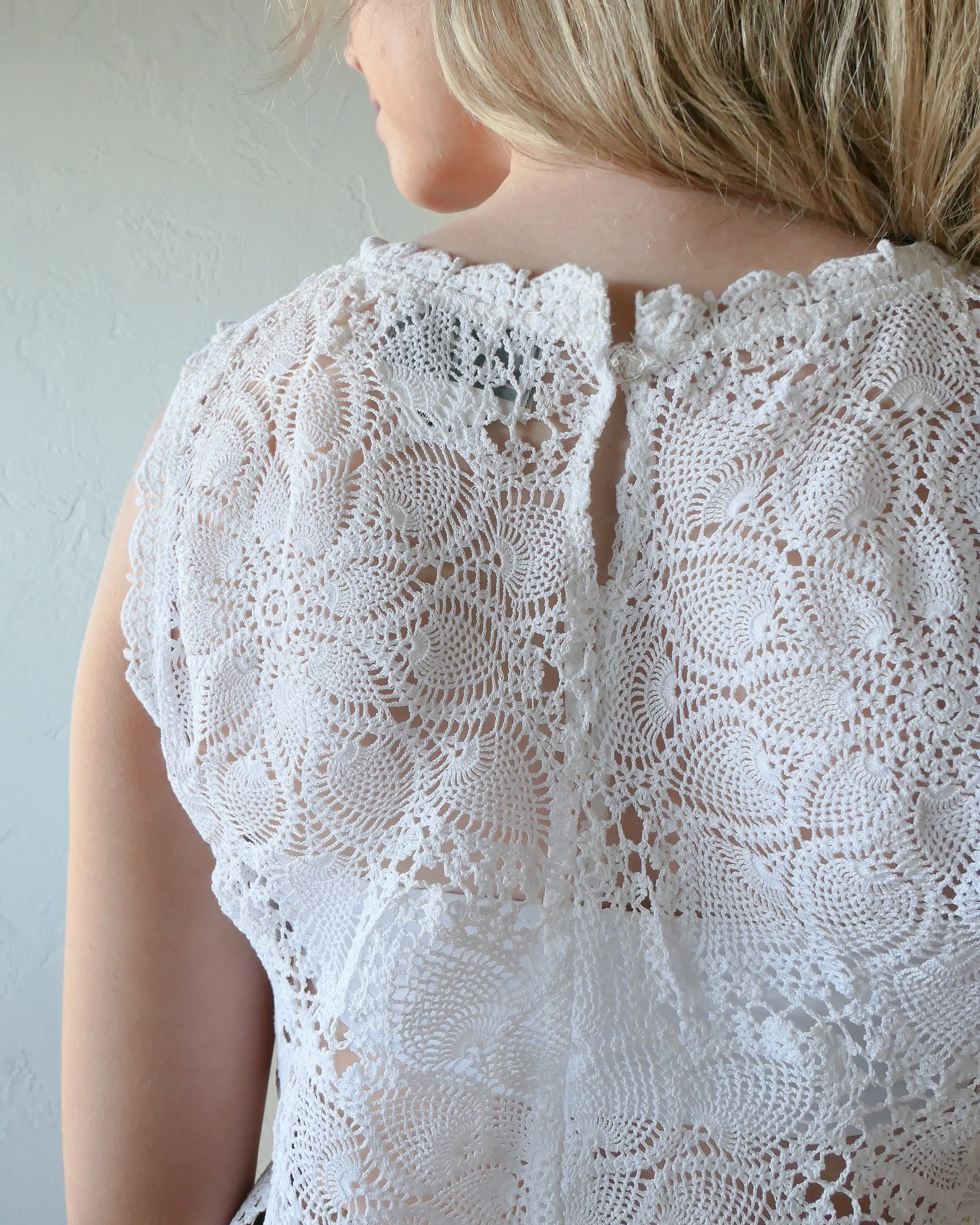 Closeup of back view of white top. An ultra casual, hip, and boho-chic crop tank top made with interconnected flower doilies reminiscent of the white lotus, which symbolizes peace, tranquility, and calmness. Lace trim at the collar and sleeve. Model is wearing a white colored crochet Lim's Vintage top.