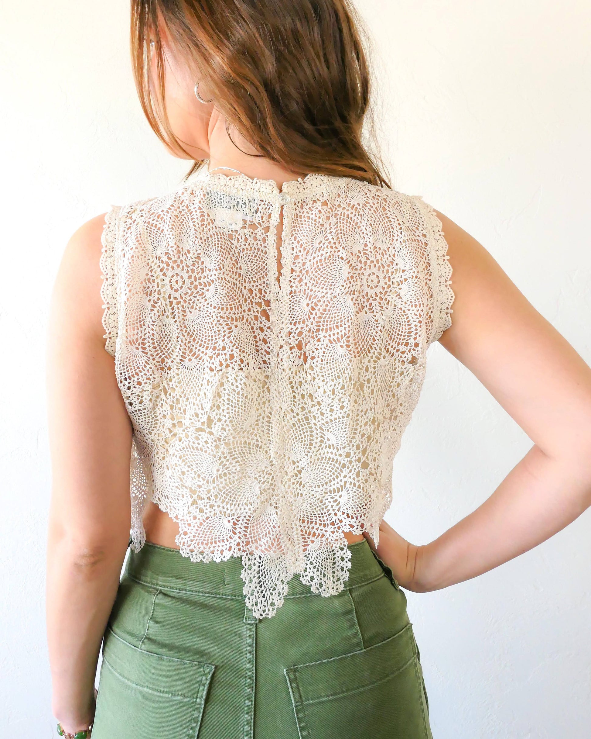 Back view of top. An ultra casual, hip, and boho-chic crop tank top made with interconnected flower doilies reminiscent of the white lotus, which symbolizes peace, tranquility, and calmness.  Lace trim at the collar and sleeve.  Model is wearing a natural colored crochet top. 