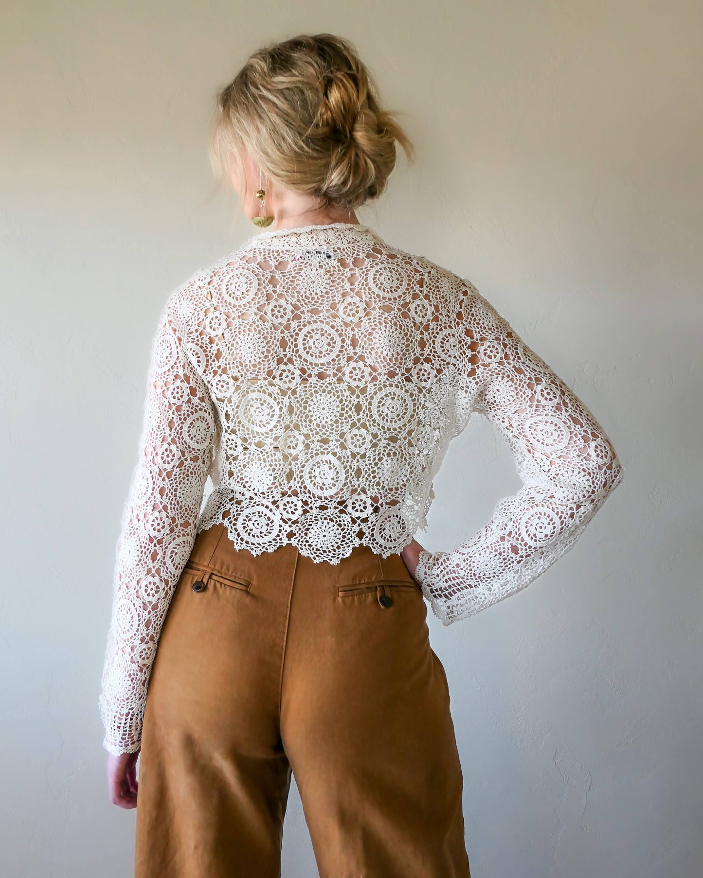 Back view of ivory colored crochet crop top. A versatile and boho chic hand crocheted cropped cardigan sweater with a unique, repeating kaleidoscope-like floral pattern throughout. Dress it down with a pair of jeans and sandals, or dress it up with neutral colored wide leg pants, heels, and a pair of vintage dangly earrings.