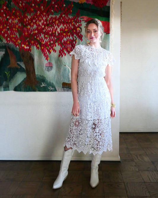 A romantic white crochet midi dress with a floral and leaf pattern, comfortable elastic waistband, and a flattering neck and shoulder design. Perfect as a vintage wedding dress or a special occasion! Wear with a white slip and heels for a refined, feminine look, or pair with boots for a more casual outing.