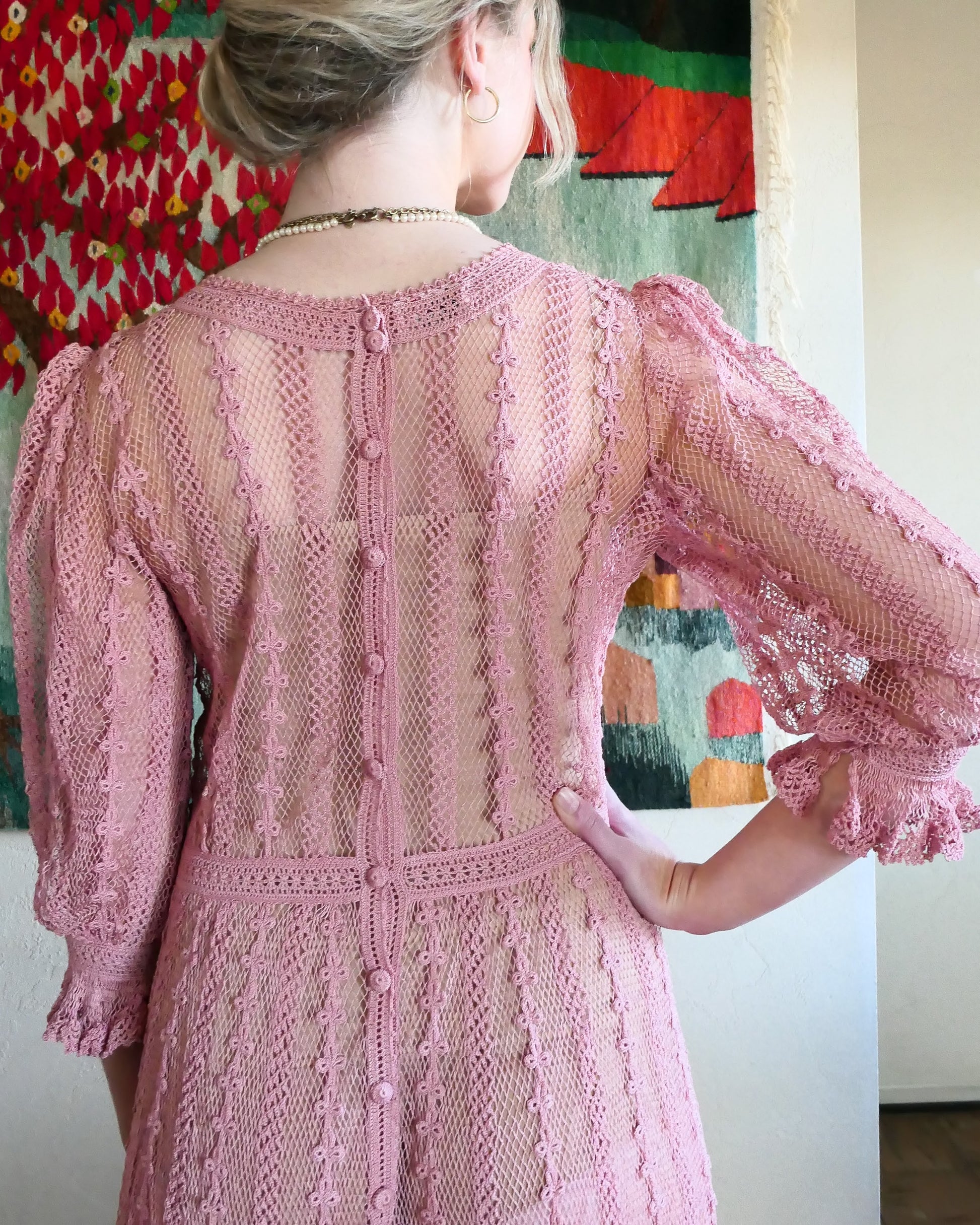 Closeup view of back of dress.  Buttons up the back.  One of our original Lim's crochet maxi dresses in a muted rose color from the 1980's. A darling of a piece and sure to turn heads, this dress was intricately hand crocheted using very fine cotton threads.   3/4 length sleeves, round collar, with ruffles at the wrist.  Model is wearing a natural colored slip underneath. 
