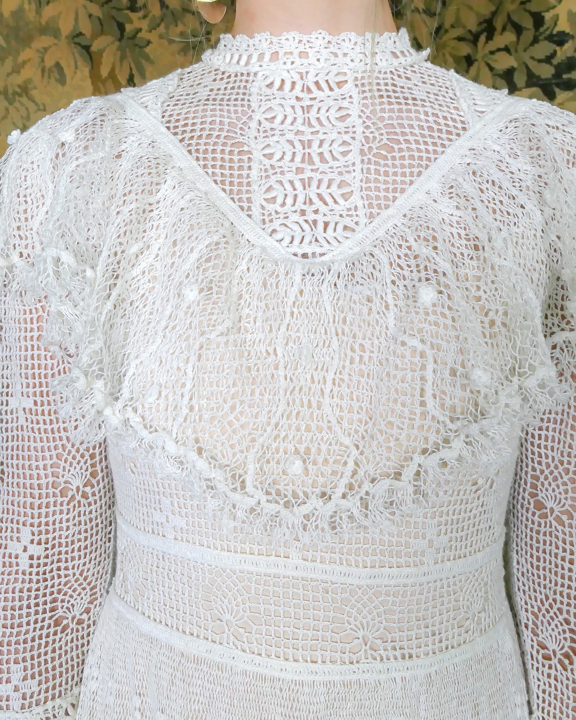 Closeup of Victorian neckline and ruffled collar. Lim's original hand crocheted maxi dress from the 1980s. Ruffle trim along the chest covering the breast area, and two tiered ruffle hem at the bottom of the dress. Very sheer crochet dress, fitted at the waist, with a tie at the back. Detailed crochet emellishment on the forearm, sleeve hem, and neckline. Model is wearing white colored dress.