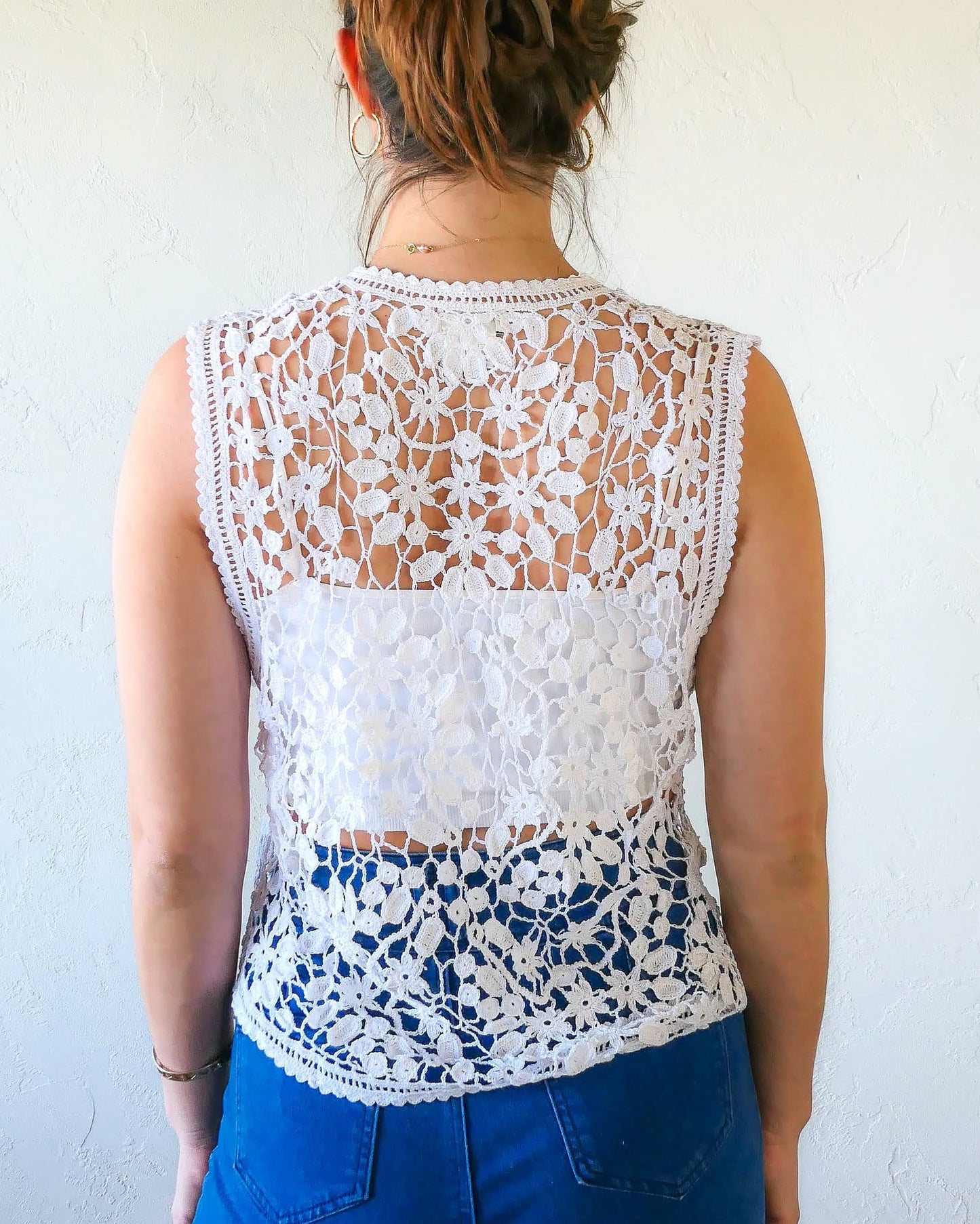Back view of vest. A Lim's Vintage original hand crocheted vest from the 1990s with a repeating jasmine floral and leaf motif throughout. Refined, delicate, and a vintage accent to your everyday wardrobe. Buttons up the front but can also be left open and worn over a bikini bra, tank top, or even a long sleeve shirt or blouse during cooler months. Due to the handmade nature of this vest, the length and bust circumference may vary slightly from piece to piece. Model is wearing white color vest.