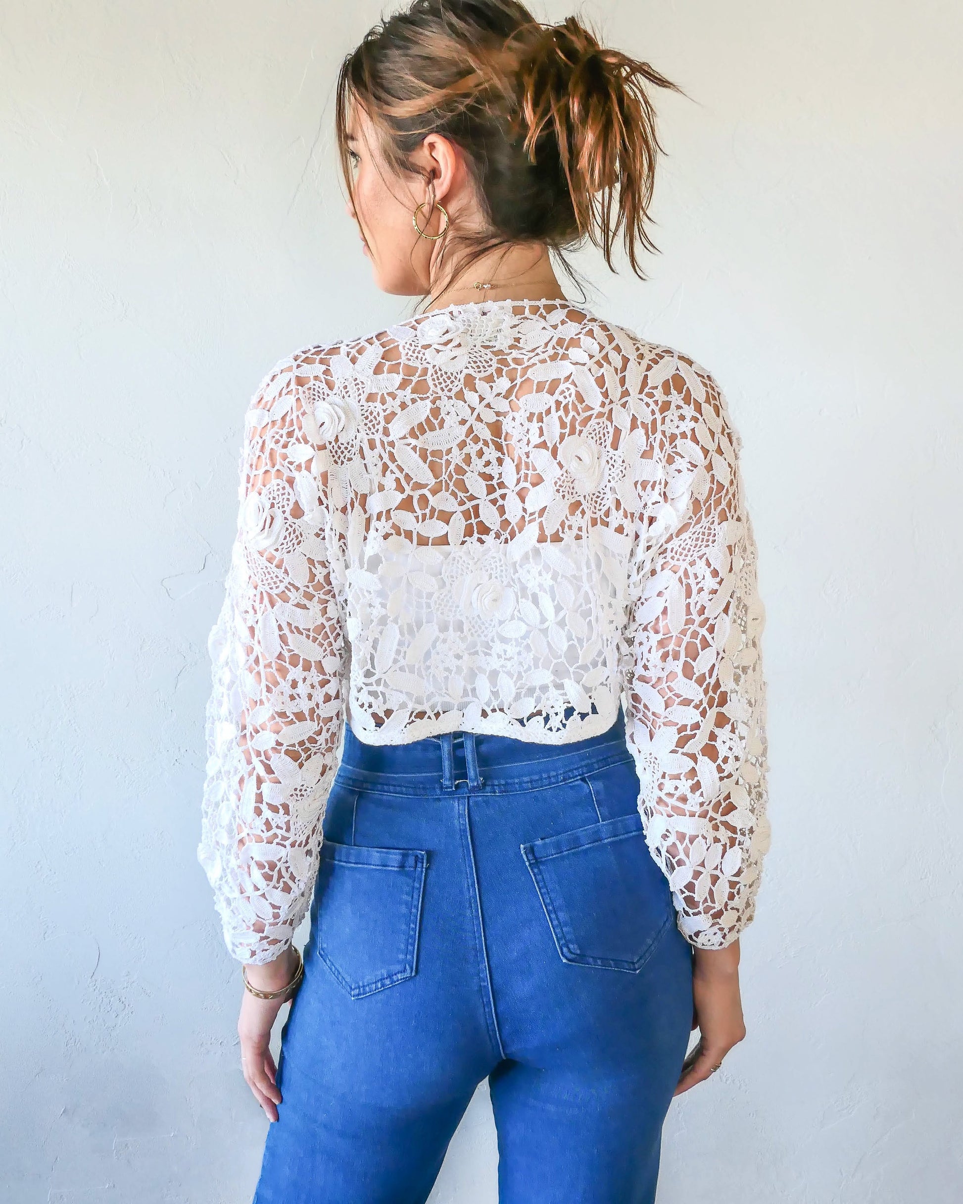 Back view. A cropped crochet long sleeve cardigan with a botanical motif of leaves and three-dimensional flowers subtly reminiscent of tea roses. Buttons ups the front, with roomy sleeves and a relaxed fit around the bust.