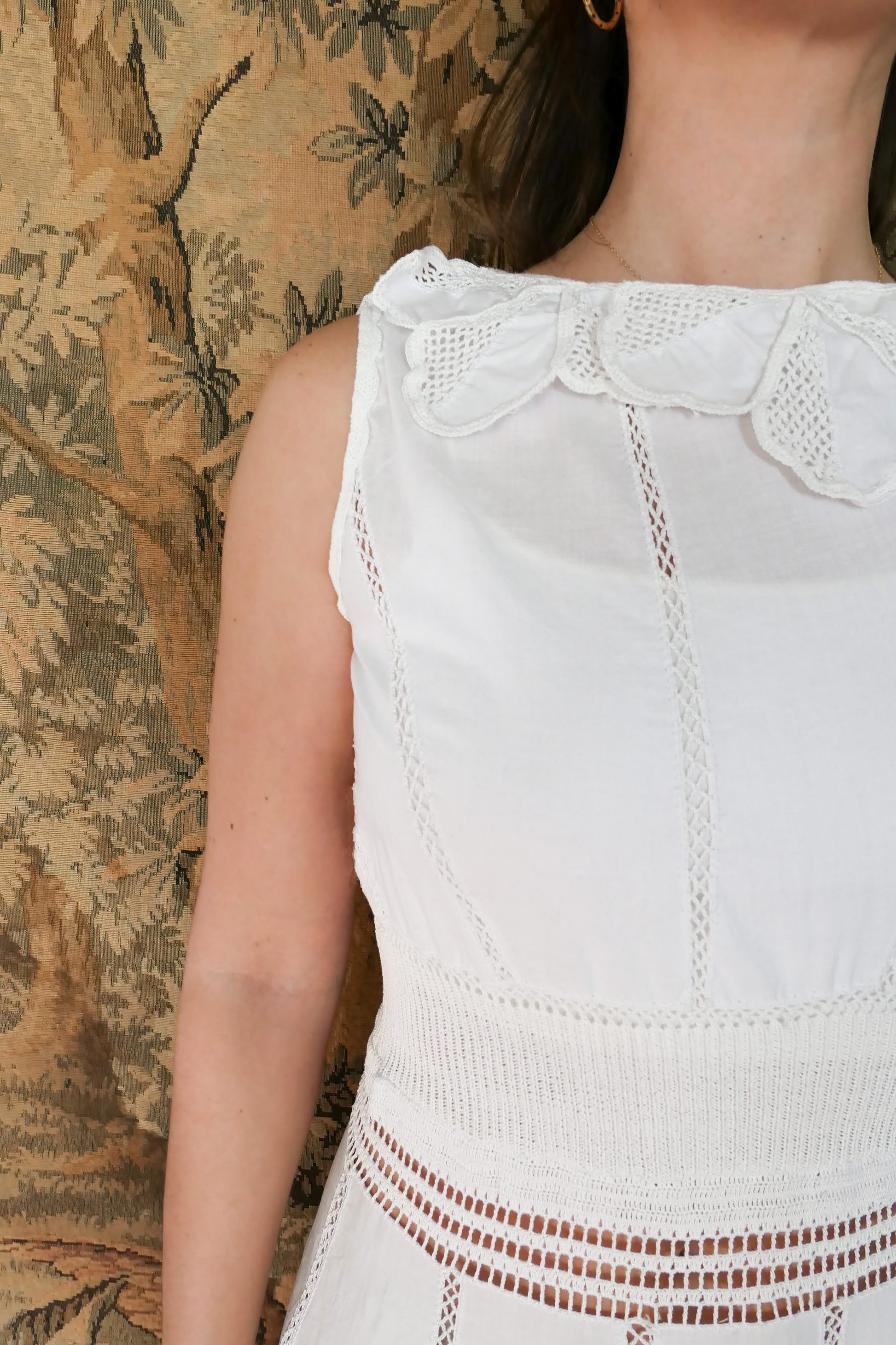 Closeup view of front of dress and collar.  A white cotton midi dress with whimsical crochet and embroidered flowers at the hem and collar, and teardrop shaped eyelet detail on the skirt. Stretchable ribbed knit fabric around the waist adds contour to the dress.
