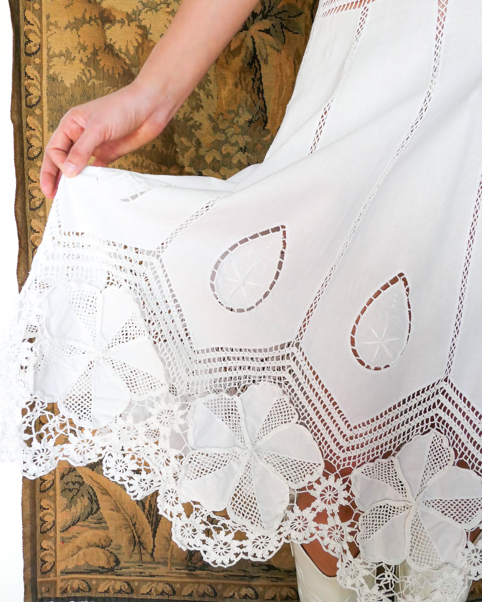 Closeup view of the hem of dress with detailed crochet and embroidery work.  A white cotton midi dress with whimsical crochet and embroidered flowers at the hem and collar, and teardrop shaped eyelet detail on the skirt. Stretchable ribbed knit fabric around the waist adds contour to the dress.