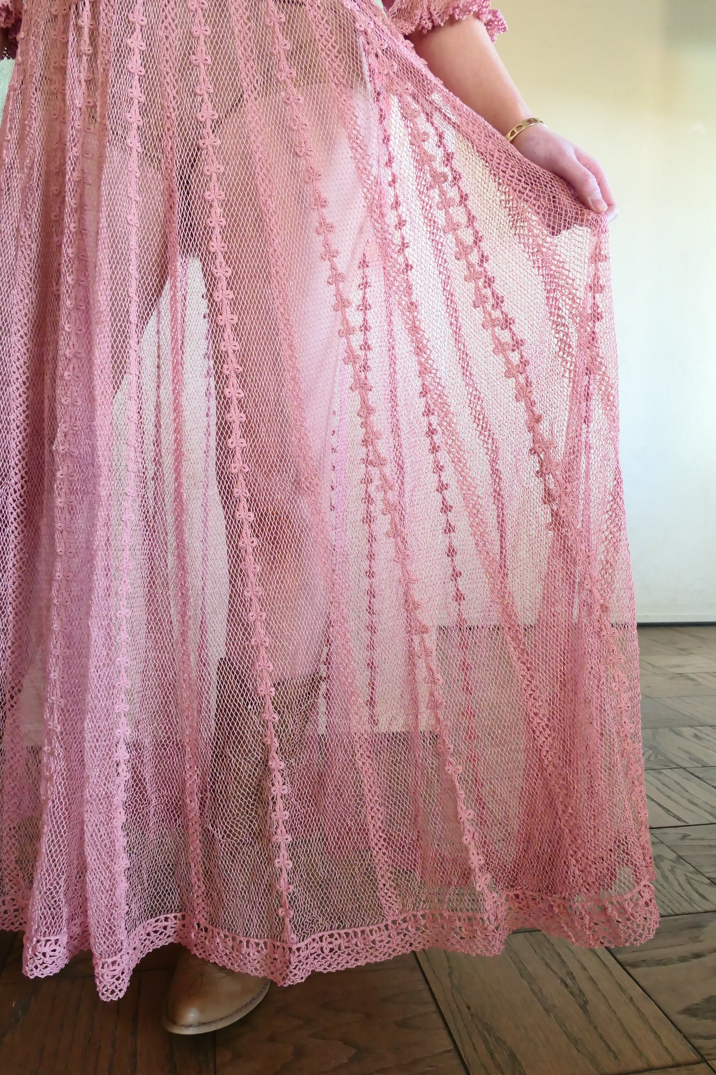 Closeup view of bottom of dress.  One of our original Lim's crochet maxi dresses in a muted rose color from the 1980's. A darling of a piece and sure to turn heads, this dress was intricately hand crocheted using very fine cotton threads.   3/4 length sleeves, round collar, with ruffles at the wrist.  Model is wearing a natural colored slip underneath. 