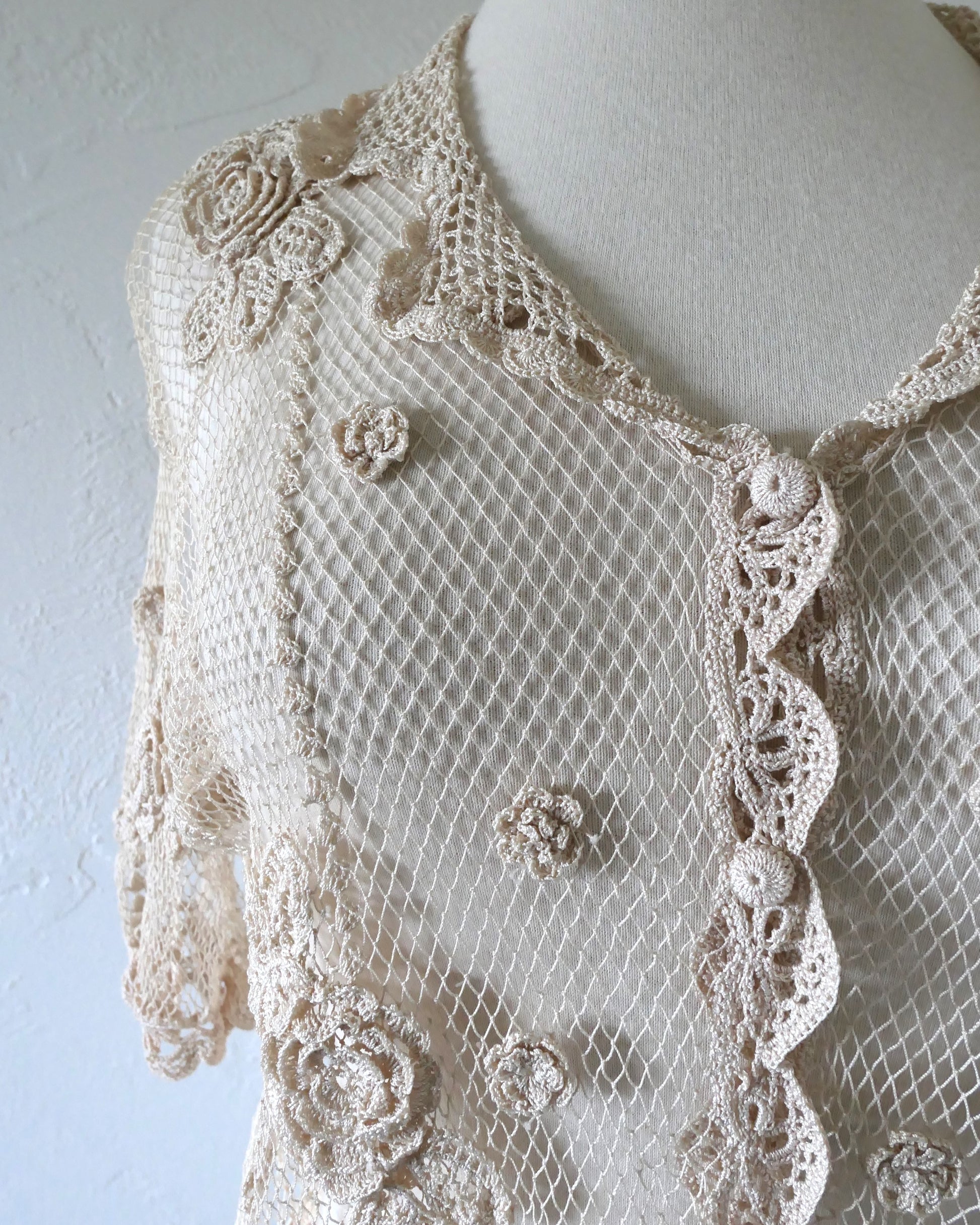 Closeup of Lim's VIntage natural colored cropped crochet cardigan with carnation floral motif, notched collar, and scalloped edging around sleeve hem and bottom hem. 