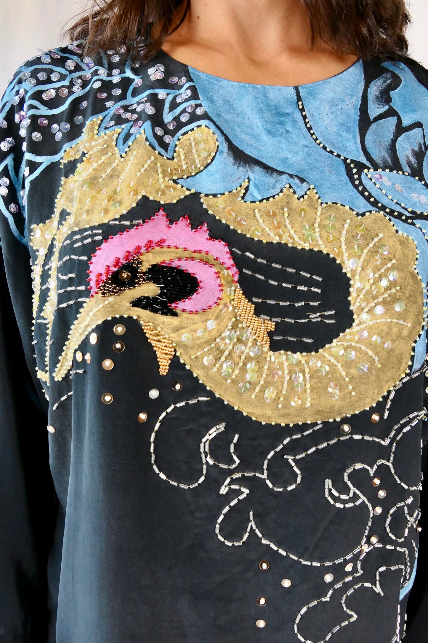 Close-up view. Make a bold party statement in this black silk pullover top with a hand-painted gold and turquoise phoenix, embellished with iridescent, pink, black, and clear colored sequins. In Chinese culture, the phoenix is a symbol of virtue, harmony, balance and peace. This beautifully made top is sure to bring harmony to your wardrobe as well!