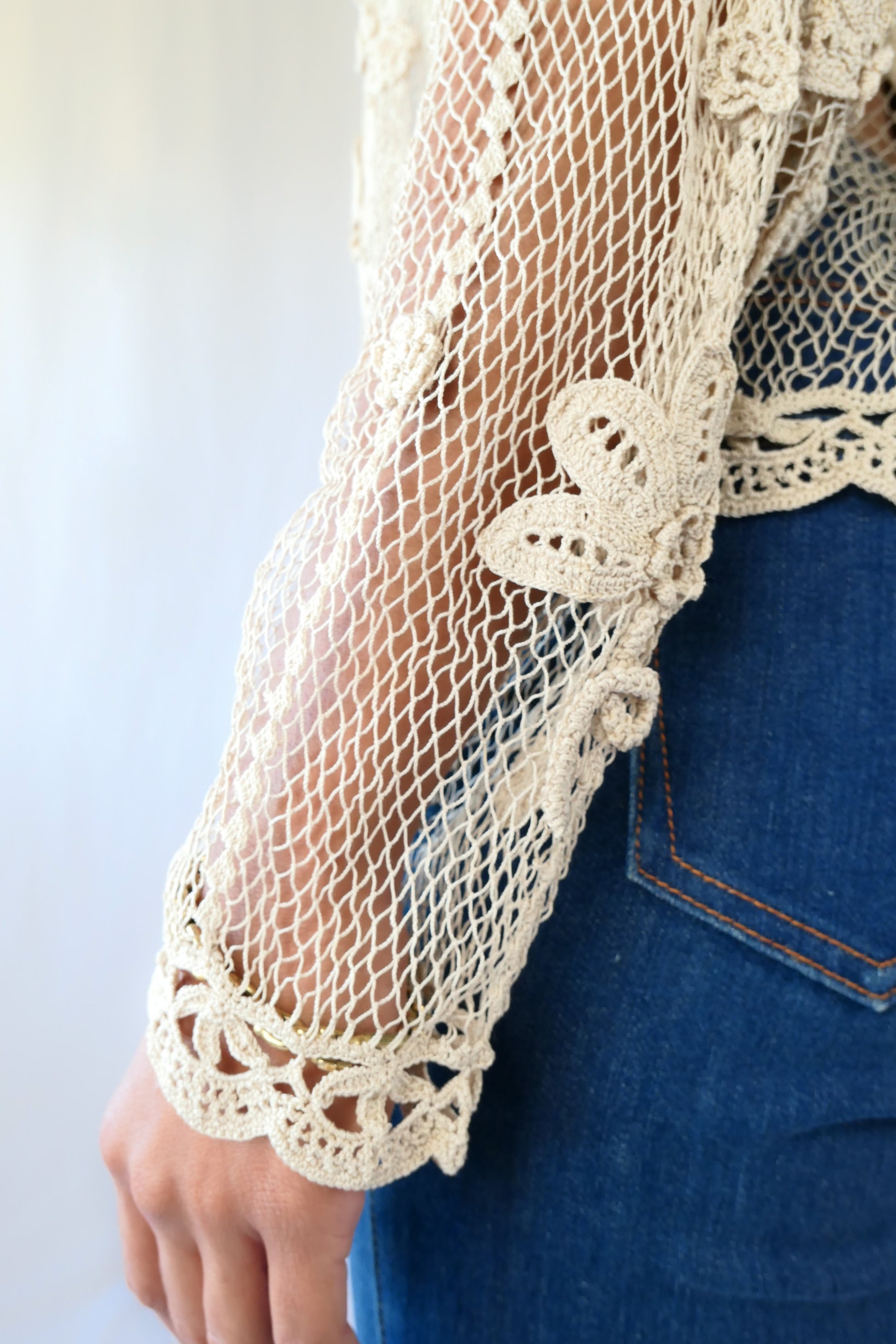 Close-up of sleeve.  A sheer, hand crocheted Lim's Vintage original long sleeve cardigan with a wide notched collar and daisy floral motif design throughout. Pair it with a tank top, jeans, and boots or sandals for a fresh, boho chic look.